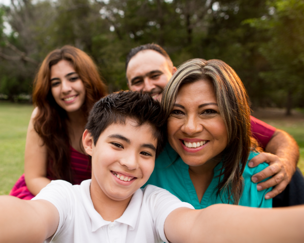 A Latinx mother, father, daughter, and son take a selfie in a park.